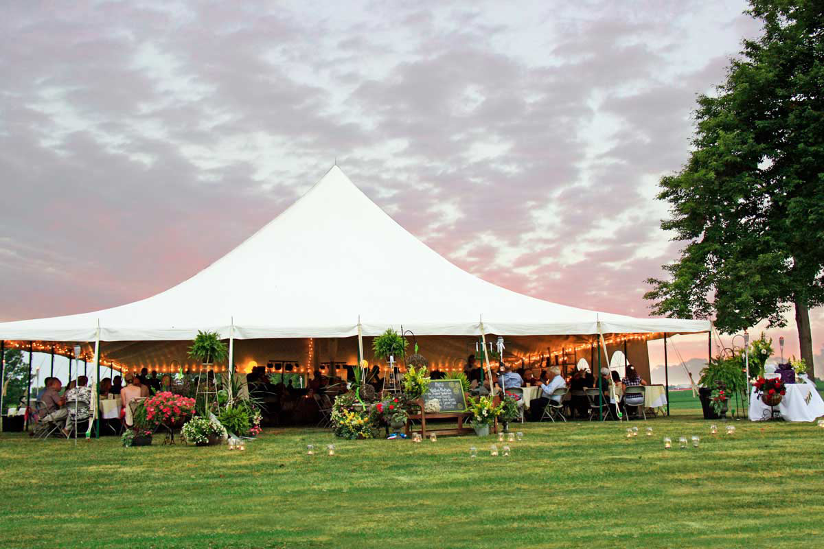 A warm pink sunset cast over a graduation party beneath a high peak tent decorated with florals